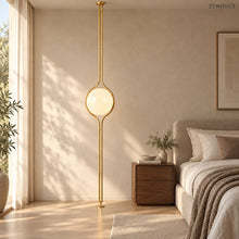 ORELIA Brass floor-to-ceiling lamp beside a linen bed in a sunlit neutral bedroom
