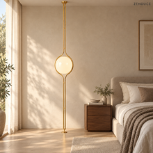 ORELIA Brass floor-to-ceiling lamp beside a linen bed in a sunlit neutral bedroom