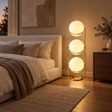 ORVENA brass three-tier globe lamp glowing beside an upholstered bed at dusk